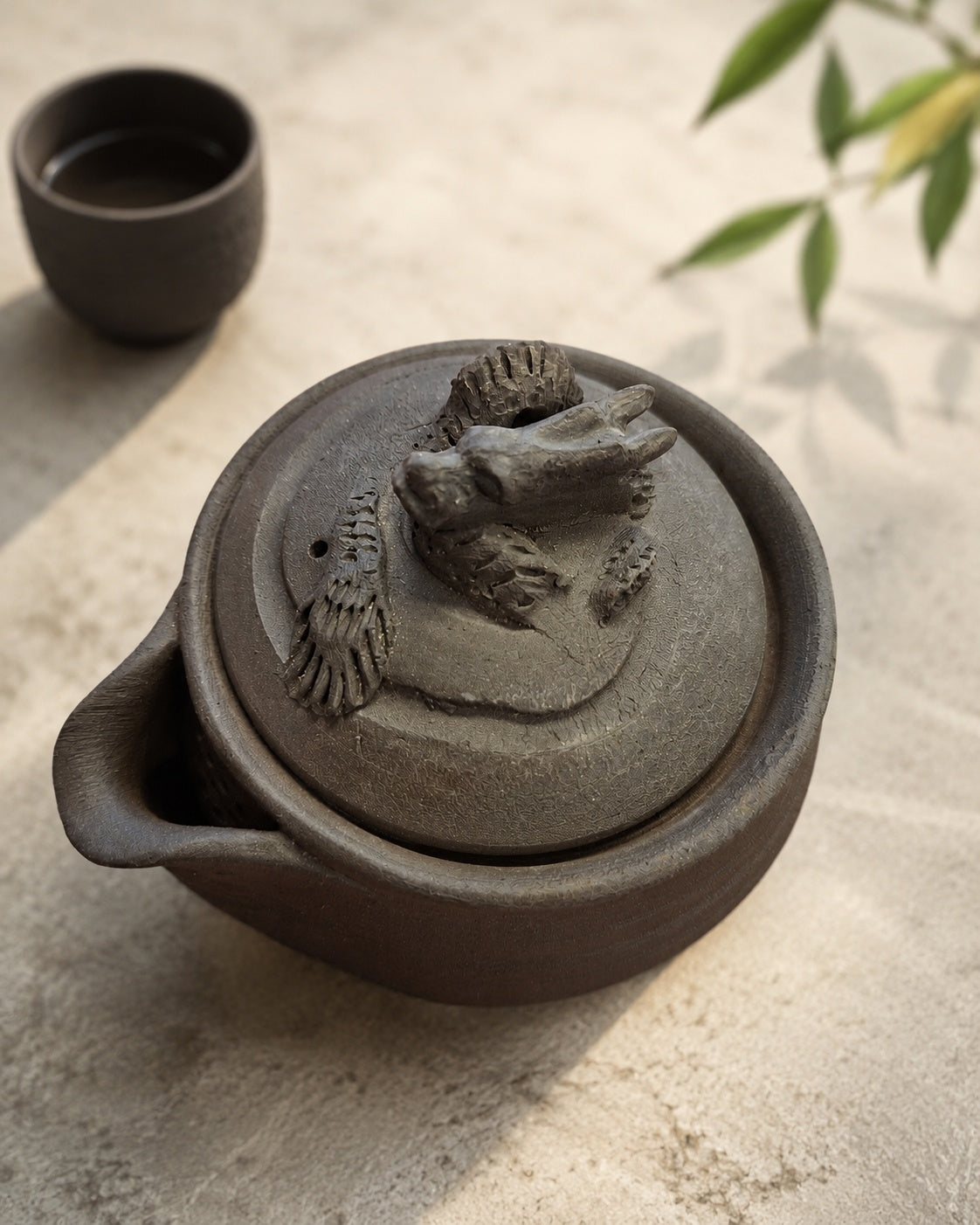 Unglazed stoneware hōhin with dragon lid on stone, teacup and green leaves behind.