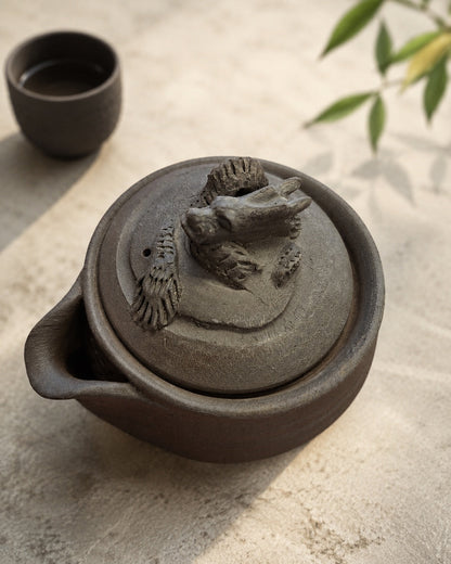 Unglazed stoneware hōhin with dragon lid on stone, teacup and green leaves behind.